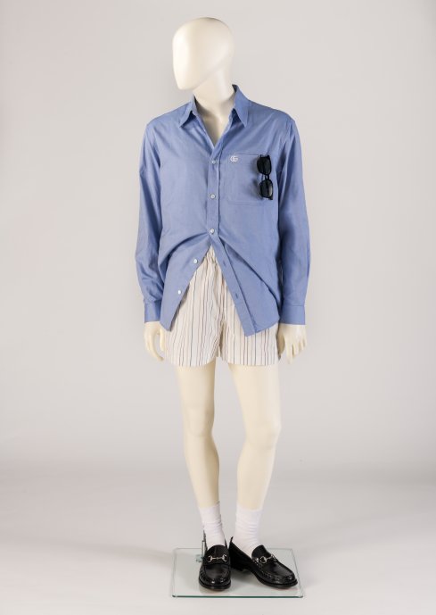 A light blue cotton shirt, striped boxer-style shorts worn with white crew socks, classic black Horsebit loafers, and sunglasses